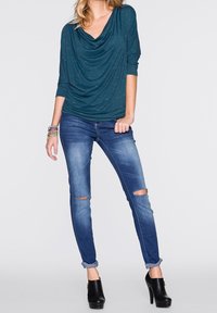 Haut drapé bleu sarcelle avec manches trois-quarts, associé à un jean skinny bleu déchiré. Bottines noires à talons hauts complètent la tenue.