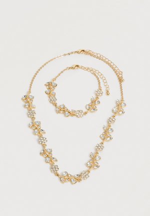 Set colier și brățară în ton auriu cu modele florale și de frunze din cristal transparent, ambele având închizători reglabile pe lanț pe un fundal alb.