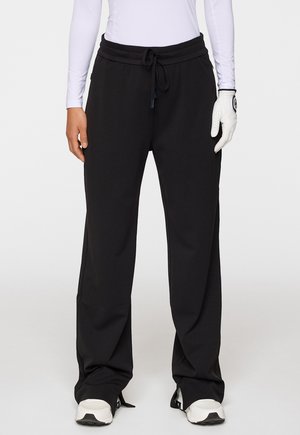 Schwarze Sporthose mit verstellbarem Bund, lockerer Passform und weichem Material. Kombiniert mit einem weißen Langarmoberteil und einem weißen Golfhandschuh.