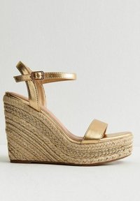 Sandalo wedge metallico oro con una larga fascia sulla punta e fibbia alla caviglia, caratterizzato da una piattaforma in corda intrecciata e superficie texturizzata.