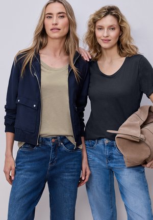 Due donne in piedi fianco a fianco; una con giacca blu scuro e top beige, l'altra con t-shirt nera che tiene una giacca in camoscio color cuoio, entrambe con jeans blu.