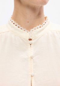 Blusa di crema con colletto rifinito in pizzo, chiusura con bottoni e tessuto strutturato. Design semplice con una finitura morbida.