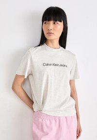 Svetlošedé tričko s krátkymi rukávmi a okrúhlym výstrihom, s čiernym logom "Calvin Klein Jeans" na prednej strane. Mäkkučký, pohodlný materiál.