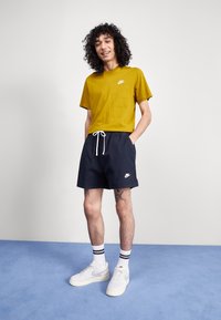 Rumena bombažna majica s logotipom Nike, mornarsko modri kratki hlače z belim vezalcem in logotipom Nike, bele tenisice in črtaste nogavice na modri podlagi.