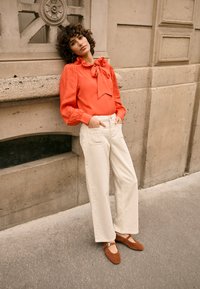 Blouse corail à manches longues avec un détail nœud, associée à un pantalon blanc taille haute à jambes larges et des chaussures en daim marron. Fond texturé.
