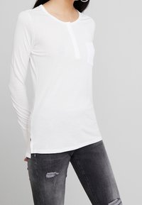 Vit långärmad skjorta i lätt tyg. Har en knäppning vid halsen, vänster bröstficka och sidslitsar. Matchas med mörka slitna jeans.