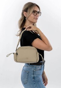 Sac à bandoulière en toile beige avec une sangle tressée, des compartiments zippés et un logo en relief. Modèle portant un haut sombre et un jean en denim.