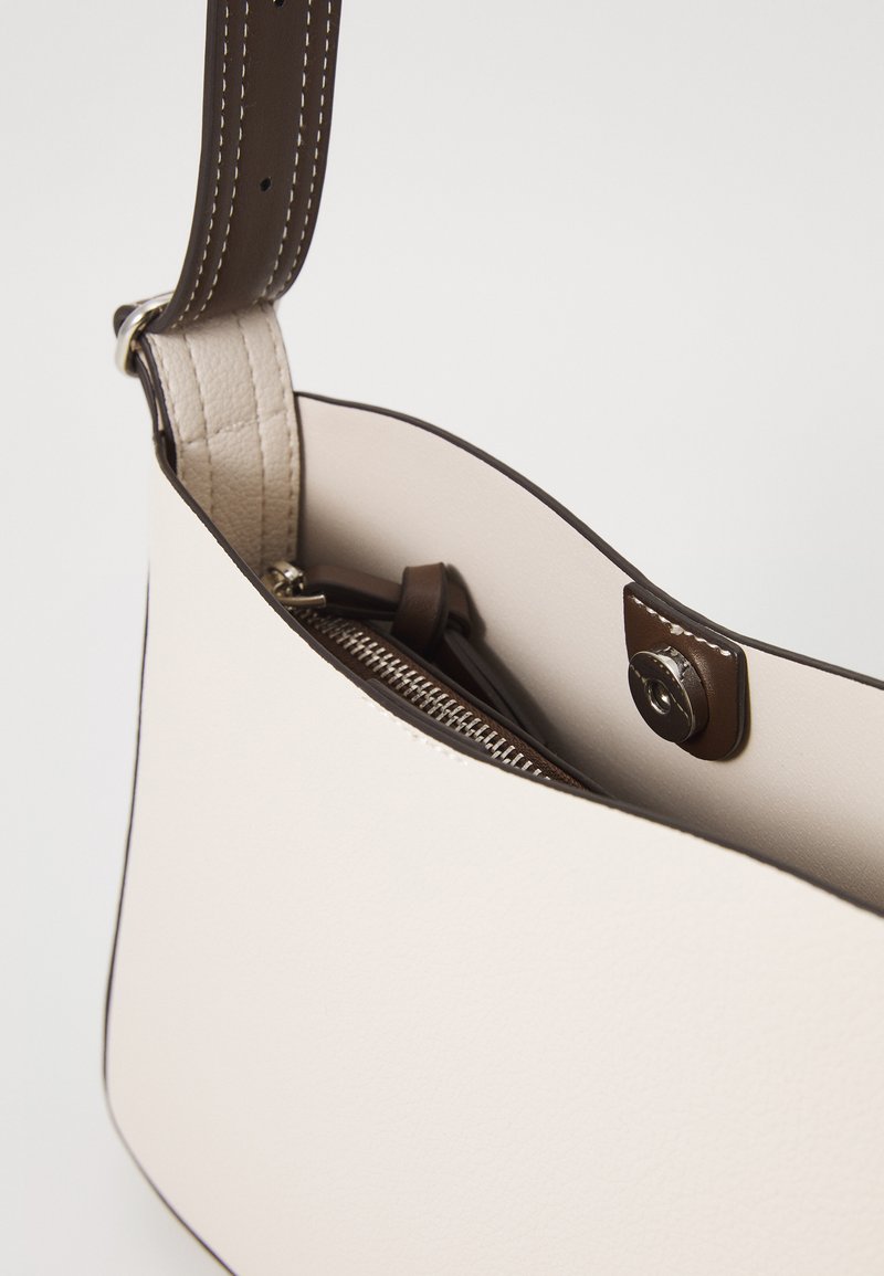 Bolso de cuero crema con una correa marrón, que presenta un cierre de cremallera y un botón a presión. Textura suave y forma sencilla y curva.