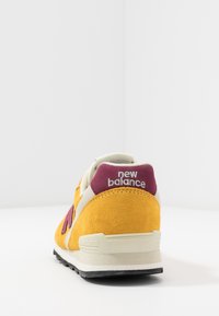 Sportskor med ovandel i gult mocka, beige detaljer, lila häl och svart gummisula. "New Balance"-logotypen syns på hälen.