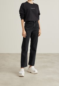 Sudadera negra de manga larga con logo, jeans negros de talle alto y zuecos blancos. El conjunto presenta un ajuste relajado, una textura suave y un diseño sencillo.