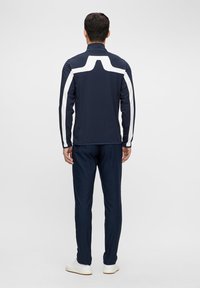 Jachetă sportivă bleu marin cu accente albe și guler înalt, purtată cu pantaloni bleumarin asortați. O țesătură fină, ușoară; design slim fit.