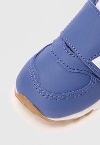 Chaussure synthétique bleue avec une bande velcro, bout arrondi et couture blanche contrastante. Présente une semelle en caoutchouc brun clair et une texture lisse.