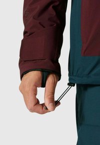 Bordeauxrote wasserdichte Jacke mit strukturierter Oberfläche. Verstellbare Bündchen und ein Kordelzugsaum sind sichtbar, mit einer Hand, die das Zugband hält.
