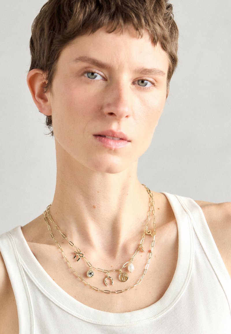 Colliers en or superposés avec divers pendentifs, comprenant une pierre, un charme et des pièces de formes variées. Les chaînes présentent un design de maillons allongés.
