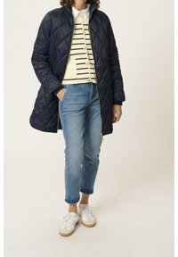 Una donna è in piedi con indosso un cappotto trapuntato blu navy sopra un maglione a righe e jeans azzurri chiari, completato da sneakers bianche.