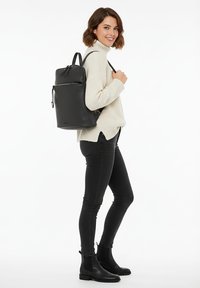 Svart läderryggsäck med en strukturerad design, tillg�ängliga fickor med dragkedja och justerbara remmar, bärs över en cremefärgad tröja och svarta byxor.