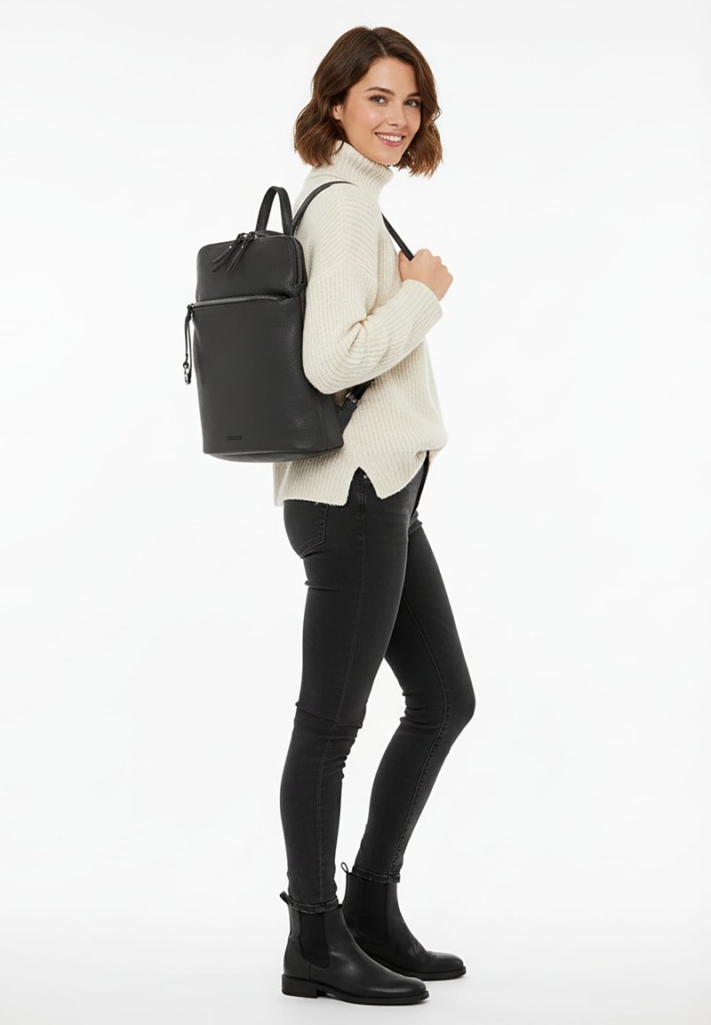 Svart läderryggsäck med en strukturerad design, tillgängliga fickor med dragkedja och justerbara remmar, bärs över en cremefärgad tröja och svarta byxor.
