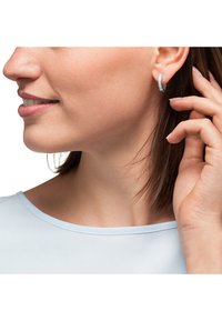 Orecchini a cerchio in argento con superficie texture, indossati all'orecchio. Il modello indossa una maglia azzurro chiaro e tocca l'orecchio con una mano.