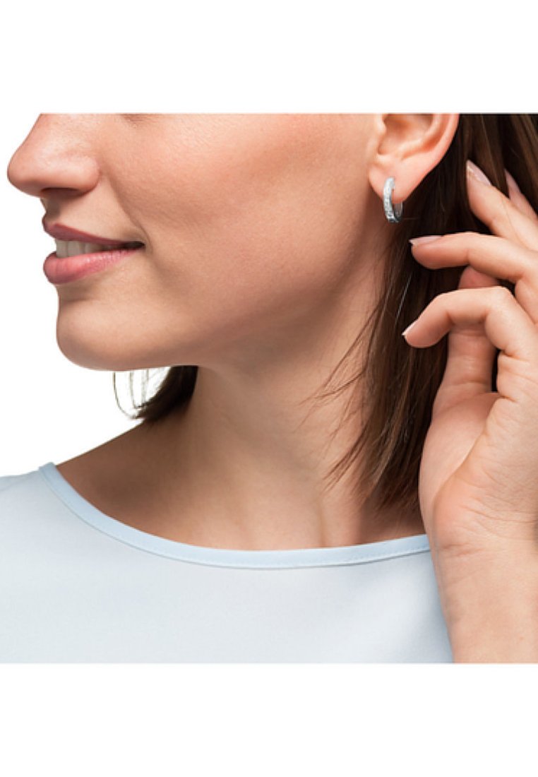 Orecchini a cerchio in argento con superficie texture, indossati all'orecchio. Il modello indossa una maglia azzurro chiaro e tocca l'orecchio con una mano.