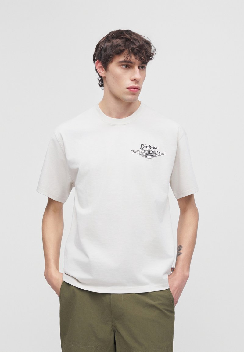 Jeune homme aux cheveux bouclés foncés portant un t-shirt blanc Dickies avec logo et un pantalon vert olive, mains dans les poches, regardant sur le côté.