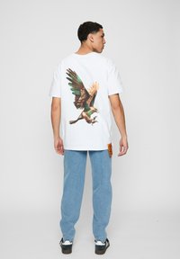 Hvid bomulds T-shirt med et farverigt ørnegraphic på bagsiden. Sammen med lyseblå denimbukser og sorte sko med hvide detaljer.