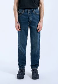 Jeans de mezclilla azul claro con corte recto, diseño de cinco bolsillos y elementos de metal bronceado, combinados con zapatillas oscuras. Se nota un ligero desgaste.
