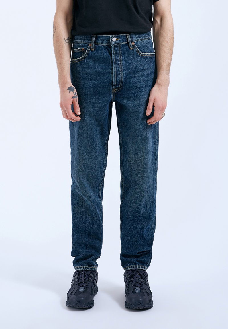 Jeans de mezclilla azul claro con corte recto, diseño de cinco bolsillos y elementos de metal bronceado, combinados con zapatillas oscuras. Se nota un ligero desgaste.