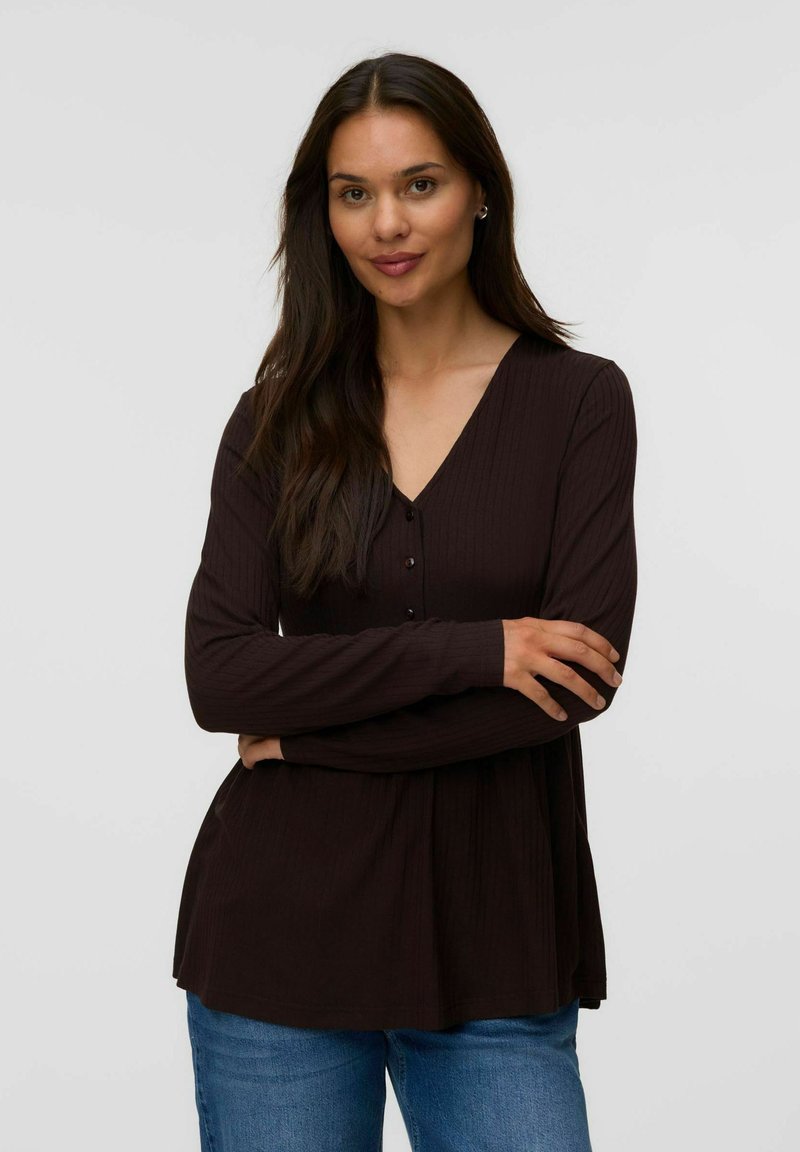 Femme aux longs cheveux foncés portant un haut noir à manches longues et col en V ainsi qu'un jean bleu, debout les bras croisés devant un fond uni.