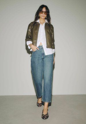 Frau trägt braune Lederjacke, weißes Hemd, blaue Jeans, flache Schuhe mit Schlangenmuster und Sonnenbrille, steht vor einem schlichten hellgrauen Hintergrund.
