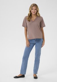 V-hals, kortærmet top i støvet rosenfarve med blonde detaljer; parret med lyseblå lige ben jeans og sorte flade sko. Glat, afslappet stof.