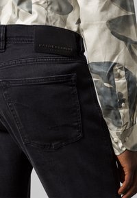 Vaqueros negros de denim con una superficie texturizada, que cuentan con un bolsillo trasero con detalle de costuras y una parche de cuero negro que dice "BALDESSARINI".