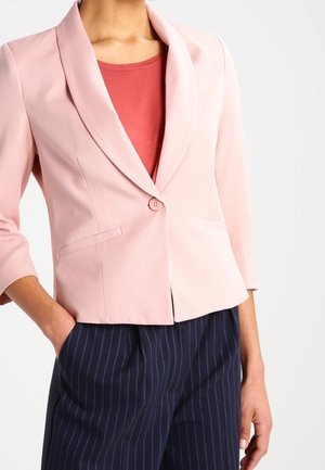 Femme portant un blazer rose clair à un seul bouton sur un haut rouge et un pantalon à rayures bleu marine, avec une main dans la poche.