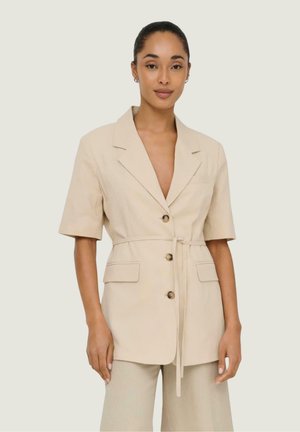 Vrouw draagt een beige korte mouw blazer met riem en knopen en bijpassende wijde broek, staand tegen een effen achtergrond.