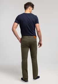 Oliwkowe spodnie o dopasowanym kroju, z tylnymi kieszeniami i brązową skórzaną metką na pasku, zestawione z granatowym t-shirtem i sportowymi butami.