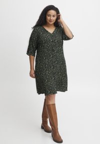 Rochie cu imprimeu floral, cu o bază neagră și accente verzi, având decolteu în V, mâneci scurte și o croială lejeră, purtată cu cizme maro până la genunchi.