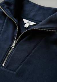 Felpa zip con colletto alto di colore blu navy, realizzata in tessuto morbido, con zip argentata e un'etichetta che mostra i dettagli del marchio e della taglia.