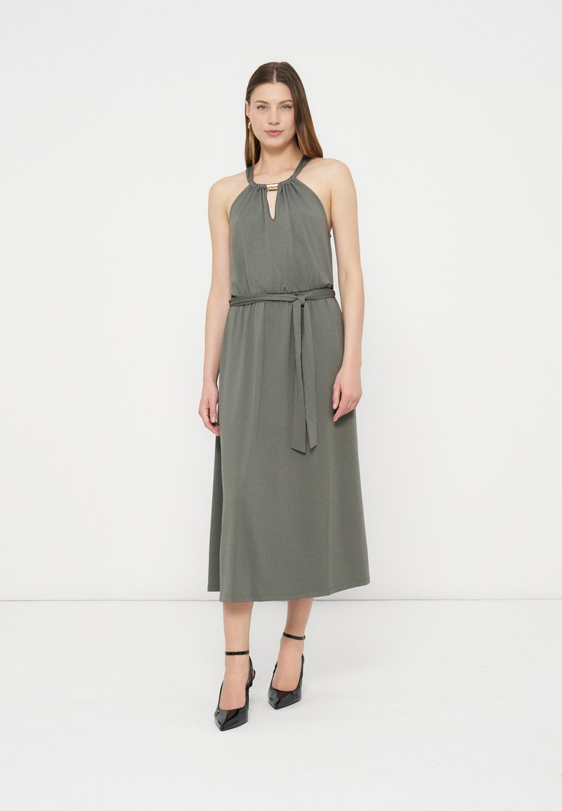 Femme portant une robe midi sans manches vert olive à encolure halter et ceinture à la taille, associée à des talons noirs pointus avec bride cheville.