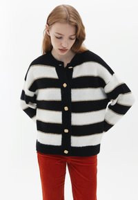 Svart och vit randig kofta med guldaccenter, med rund hals, stora träknappar och en mjuk, stickad textur.