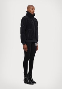 Svart fleecejacka med hög krage, dragkedja fram och strukturerade paneler, kombinerad med svarta leggings och ankelstövlar.