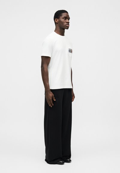 Λευκή βαμβακερή μπλούζα με τσέπη που έχει πολύχρωμο γραφικό σχέδιο, σε συνδυασμό με φαρδιά μαύρη παντελόνια και μαύρα παπούτσια.