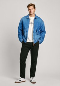 Chaqueta de denim, azul claro, con cierre frontal y dos bolsillos laterales. Camiseta blanca debajo, pantalones negros y zapatillas blancas con detalles.