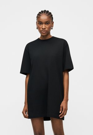 Mladá žena s vrkočmi, ktorá nosí zlaté kruhové náušnice a nadrozmerné jednoduché čierne tričkové šaty, stojaca na bielom pozadí.