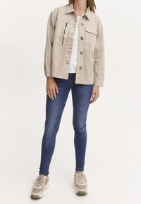 Jasnobeżowa kurtka dżinsowa z zamykaną na zamek kieszenią na piersi, zapinana na guziki, o luźnym kroju, zestawiona z niebieskimi obcisłymi jeansami i trampkami.