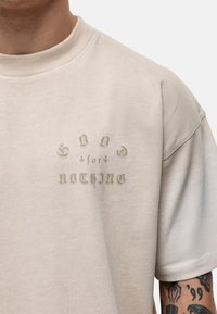 Kremowa, oversize'owa koszulka z miękkiego materiału, z vintage'owym nadrukiem w stonowanym brązie umieszczonym na środku klatki piersiowej.