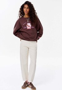 Žena s dlouhými kudrnatými vlasy, oblečená v hnědém svetru s růžovým grafickým motivem psa, sedí na křesle. Má na sobě béžové kalhoty a hnědé tenisky s růžovými ponožkami.