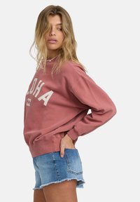 Ružová mikina s nápisom "ALOHA" v bielej farbe, s voľným strihom, okrúhlym výstrihom a rebrovanými manžetami, kombinovaná s džínsovými šortkami.