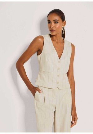 Frau mit dunklen Haaren in einer ärmellosen, beigefarbenen Weste mit Nadelstreifen und passender Hose, die mit einer Hand in der Tasche vor einem schlichten Hintergrund steht.