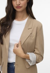 Blazer beige con rever a punta, chiusura a un bottone e taschino sul petto, abbinato a una camicia bianca e polsini a righe arrotolati.