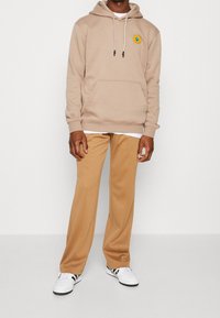 Person som bär en beige hoodie med en gul cirkelformad tygmärke, kamelfärgade byxor och vita sneakers med svarta ränder, stående mot en enfärgad bakgrund.