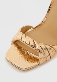 Χρυσό μεταλλικό πέδιλο με ψηλό τακούνι, τετράγωνη μύτη, διαπλεκόμενα λουράκια με υφή που μοιάζει με ερπετό και λείο εσωτερικό ταίριασμα.